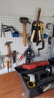 Wide view of pegboard wall with various hanging hand tools including hammers, pliers, saw, and a plastic toolbox on the workbench below.