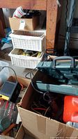 Box with multiple tools including a gas can, white woven storage baskets containing various supplies, and other assorted tools and electrical items.
