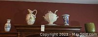Wide view of five ceramic items on wooden furniture against a red wall including two vases, one tureen, one pitcher, and one planter.
