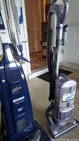Photo of two vacuums: blue Kenmore Progressive and gray Shark Navigator Lift-Away, shown upright side by side.