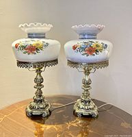Pair of 23-inch brass hurricane lamps with hand painted milk glass floral shades shown side by side on wooden table.