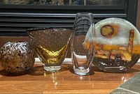 Four modernist blown glass art pieces displayed on wood surface by a window. Includes two Sommerso bubble vases, a clear tall vase, and a fused glass plate with abstract design.