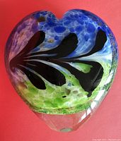 Close-up front view of the heart-shaped glass paperweight on red background showing blue, black, green and clear glass Murrini design.