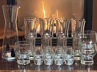 Full set of clear glass barware displayed with fireplace background including 8 pilsner glasses, 8 shot glasses and 1 carafe pitcher.