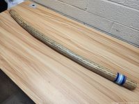 Full view of the rain stick, curved and with natural wood grain pattern. Blue and white rings near the end.
