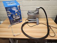 Photo of the Conair fabric steamer unit and original product box on a wooden table against a white brick wall background. The unit is silver and black with a flexible hose and detachable handheld steamer head.