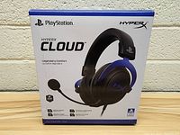 Front view of box showing HyperX Cloud headphones with PlayStation branding, listing key features such as legendary comfort, stereo drivers, memory foam, premium leatherette, aluminum frame, and 2-year warranty.