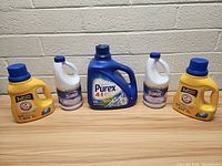 Photo showing all five bottles of laundry cleaning products aligned on wooden surface against a white brick wall background: 2 yellow Arm & Hammer detergents, 2 white Clorox bleaches, and 1 large blue Purex detergent.