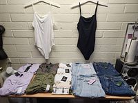 Lot of assorted new women's clothing items displayed on table and hanging on hangers against a brick wall.