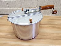 Side angled view of the aluminum stovetop popcorn maker showing the pot, lid with stirring mechanism, and wooden handle with crank.