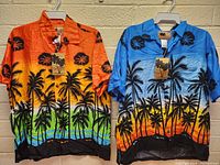 Pair of men's Hawaiian shirts hanging on white plastic hangers against a brick wall. One shirt is orange with a tropical sunset and palm tree print, the other shirt is blue with similar design.