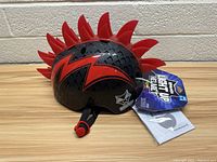 Side profile of black and red Raskulls light-up helmet with red spikes attached along the top. Tag showing "Light Up Helmet" with size L 58-60 cm attached. Red lightning bolt design on the side, ventilation holes throughout helmet surface.