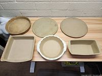 Top-down view of six stoneware baking items on wooden table against brick wall, showing different shapes and sizes.