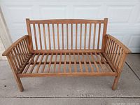 Front-facing view of a wooden slatted bench showing the backrest, arms, and seat slats, placed on a concrete surface.