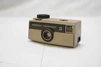 Closed front view of Kodak Hawkeye Instamatic II camera in warm gray with black lens detail and viewfinder, showing branding and model name.