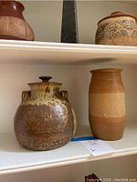 Two pieces of art pottery on a white shelf, showing the lidded jar with handles and cylindrical vase side by side.