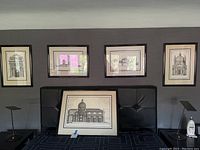 Five framed prints depicting Italian architecture hung on a dark gray wall above black tufted furniture, showing variation in size and artist signatures.