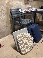 Two dark gray outdoor plastic chairs with four assorted cushions: beige patterned, gray striped, and two solid dark blue cushions, shown in basement against stone wall.