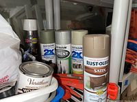 Photo showing several Rust-Oleum spray paint cans including Ultra Cover 2X and Satin finishes, and Varathane wood finish can, grouped on a shelf with some other items
