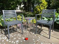 Two grey resin chairs and one folding metal table with perforated top, shown placed on a patio with plants in the background.