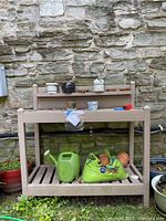 Full view of beige resin potting table with various ceramic pots on upper shelf, watering can, soil bag, and small terra cotta pots on bottom shelf. Positioned against stone wall outdoors.