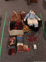 Shows all items in the lot arranged on the floor including CDs, glass dish with poinsettia design, stuffed snowman, wreath, small woven basket, and Christmas tree ornament.