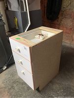 Small cupboard with three drawers, white fronts, and round knobs shown from an angled side view in basement area.