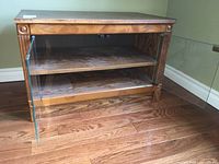 Full view of the entertainment unit showing wood grain laminate, glass doors open to reveal two shelves inside, and overall condition on hardwood floor against green wall.