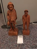 Photo showing two wooden folk art carvings on the floor: one larger man with fish net and fish, signed Turcotte, and a smaller man with a cane, signed by another artist.