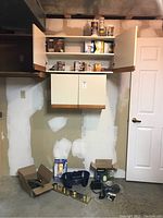 Wide view of the wall with two wooden cabinets mounted, showing some items on shelves inside cabinets and an axe, cleaning caddy, power strip and other items on the floor.