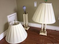 Two cream-colored table lamps on floor, both with pleated fabric shades, brass/gold-tone bases and supporting columns, approx. 27 inches tall. One lamp has visible socket damage.