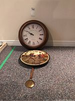 Photo showing two wall clocks placed side by side on the floor: circular Edinburgh Clerk Works Co. clock on the left and the oval St Ives Golf Club pendulum clock on the right.