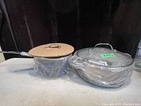 Two brand new Gotham pots in original plastic wrap, one smaller pot with long handle and one larger steamer pot with two side handles and glass lid