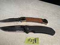 Photo of two folding knives side-by-side on a surface, one with wood and brass handle and the other black Husky knife with partial serration.