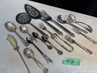 Overall photo showing seven antique and vintage silver plated cutlery and serving pieces laid on a flat surface with a label #16