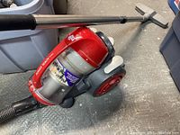 Red Dirt Devil Power Reach canister vacuum cleaner with hose lying on a rough textured floor, showing wheels and transparent dust container.