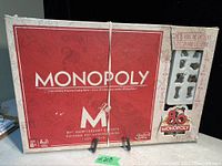 Photo of the Monopoly 80th Anniversary Edition board game box with visible wear and a plastic tray holding eight tokens visible through a window.