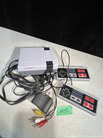 Front side view of the Nintendo Entertainment System console with two wired controllers and AV cables visible, placed on a surface against a black background.
