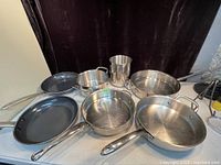 Seven pots and pans on display including Wolfgang Puck stainless steel pots, Blue Diamond frying pans with blue interior, and one KitchenAid stainless steel pan