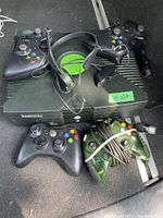 Photo shows one black Original Xbox console with the disc tray door open. Five Xbox controllers are placed around the console, including three black and two translucent green. A wired headset with microphone and the power cord are also visible.