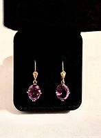Front view of the 14K gold leverback earrings with faceted oval amethyst gemstones set in gold prongs against a black jewelry box background.