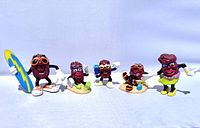 Group shot of 5 beach themed California Raisins figurines standing in a row against white background, showing variety of poses and accessories.