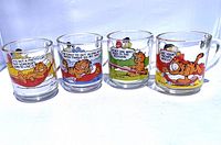 Front view of four clear glass Garfield mugs showing colorful cartoon scenes and sayings on each.