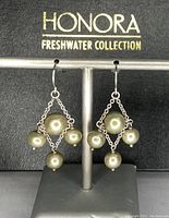 Front view of Honora earrings showing diamond-shaped chain design and pale olive green pearls on display stand with Honora Freshwater Collection box in background.
