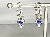 Front view of the pair of Judith Ripka earrings hanging on a display stand showing the blue sapphire center stones surrounded by cubic zirconia accents in sterling silver twisted rope design hoops.