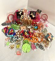 Wide view of colorful costume jewelry including various beaded necklaces, bracelets, and earrings spread out on a white surface with a pink display box behind