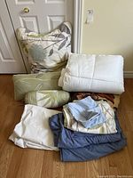 Photo shows assortment of pillows, bolster pillow, folded sheets and white comforter or mattress pad against a door.