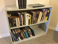 Small light gray bookshelf with multiple books on two shelves; top surface holding additional books and boxed encyclopedia volumes.
