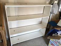 Front view of the white particle board and melamine bookcase showing three fixed shelves with dust and some sagging.