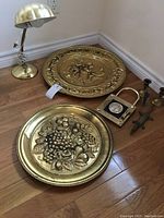 Overview of the lot showing two brass platters, thermometer in lock frame, brass lamp, and candle holders arranged on hardwood floor.